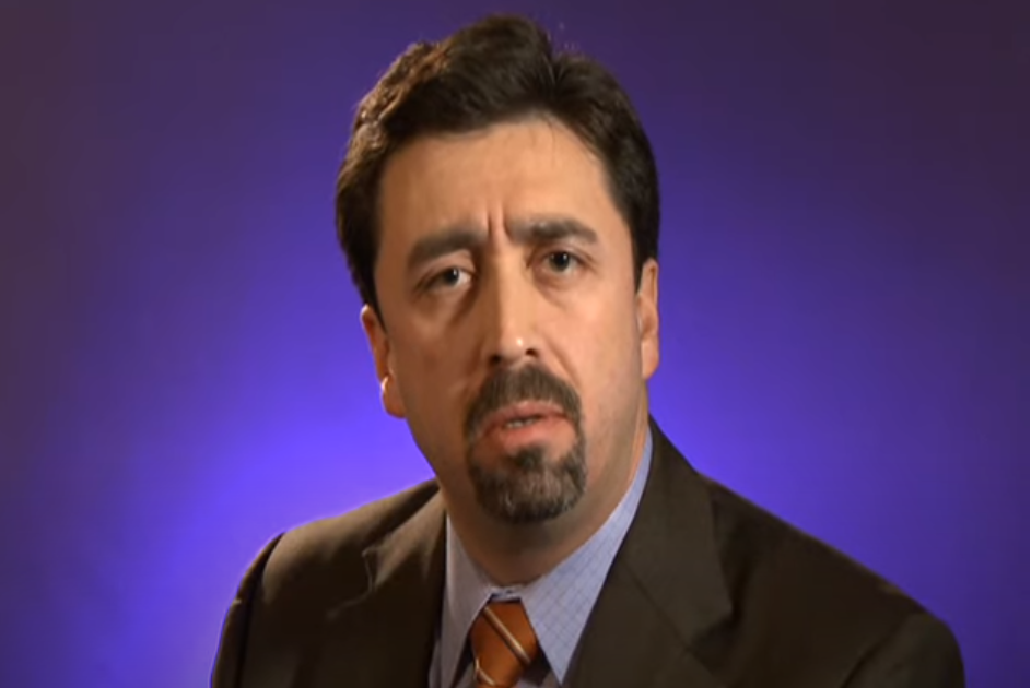 A light-skinned man with dark hair and a beard talks in front of a blue screen.
